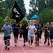 A group of military personnel and civilians are running together on a red outdoor track, with one participant carrying a POW/MIA flag. The event appears to be a commemorative run or memorial event honoring prisoners of war and those missing in action. The setting includes trees, a playground structure in the background, and overcast skies suggesting light rain.