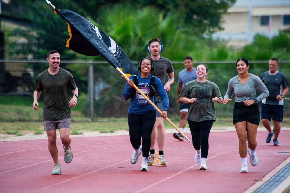 A group of people in athletic/military-style clothing are running together on a red track. A woman in the center is carrying a large black flag with gold fringe, and several participants are wearing headphones. The group appears to be participating in a commemorative or unit run event, likely on a military installation based on the attire and flag.