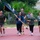 A group of people in athletic/military-style clothing are running together on a red track. A woman in the center is carrying a large black flag with gold fringe, and several participants are wearing headphones. The group appears to be participating in a commemorative or unit run event, likely on a military installation based on the attire and flag.
