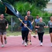 A group of people in athletic/military-style clothing are running together on a red track. A woman in the center is carrying a large black flag with gold fringe, and several participants are wearing headphones. The group appears to be participating in a commemorative or unit run event, likely on a military installation based on the attire and flag.