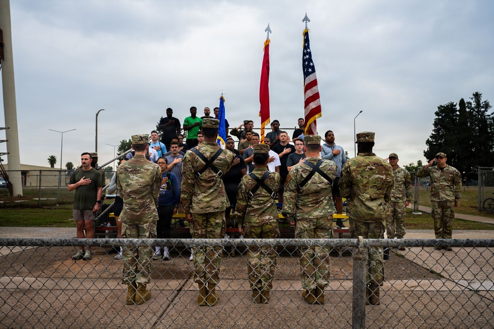 A group of military personnel and civilians are gathered outdoors during what appears to be a formal ceremony or formation, with hands placed over hearts in a pledge or national anthem observance. Three flags are prominently displayed, including the United States flag, a red military unit flag, and a blue Air Force flag. The scene takes place near a chain-link fence at what appears to be a military installation.