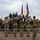 A group of military personnel and civilians are gathered outdoors during what appears to be a formal ceremony or formation, with hands placed over hearts in a pledge or national anthem observance. Three flags are prominently displayed, including the United States flag, a red military unit flag, and a blue Air Force flag. The scene takes place near a chain-link fence at what appears to be a military installation.