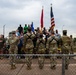 A group of military personnel and civilians are gathered outdoors during what appears to be a formal ceremony or formation, with hands placed over hearts in a pledge or national anthem observance. Three flags are prominently displayed, including the United States flag, a red military unit flag, and a blue Air Force flag. The scene takes place near a chain-link fence at what appears to be a military installation.