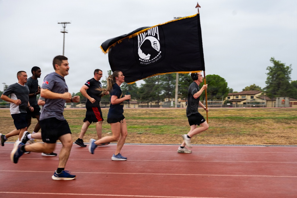 A group of military personnel or service members are running on a red track while one participant carries a POW/MIA flag with gold fringe on a wooden staff. The event appears to be a memorial or commemorative run honoring prisoners of war and those missing in action. The setting is an outdoor athletic track at what appears to be a military installation.