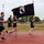A group of military personnel or service members are running on a red track while one participant carries a POW/MIA flag with gold fringe on a wooden staff. The event appears to be a memorial or commemorative run honoring prisoners of war and those missing in action. The setting is an outdoor athletic track at what appears to be a military installation.