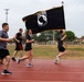 A group of military personnel or service members are running on a red track while one participant carries a POW/MIA flag with gold fringe on a wooden staff. The event appears to be a memorial or commemorative run honoring prisoners of war and those missing in action. The setting is an outdoor athletic track at what appears to be a military installation.