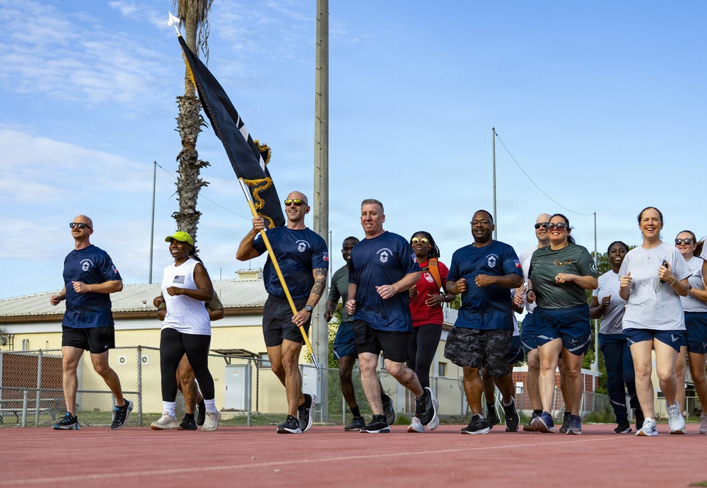 A group of military personnel and civilians are running together on an outdoor track, with one individual carrying a dark blue military guidon/unit flag. The participants are wearing a mix of navy blue military PT (physical training) shirts, civilian athletic wear, and military uniforms. The event appears to be an organized unit run or physical fitness formation event at a military installation.