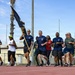 A group of military personnel and civilians are running together on an outdoor track, with one individual carrying a dark blue military guidon/unit flag. The participants are wearing a mix of navy blue military PT (physical training) shirts, civilian athletic wear, and military uniforms. The event appears to be an organized unit run or physical fitness formation event at a military installation.