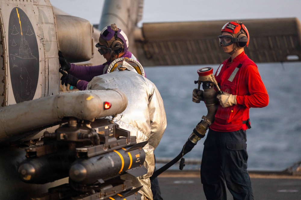 Two military personnel are conducting aircraft servicing operations on what appears to be a naval vessel flight deck over open water. One crew member in a silver/reflective fire-resistant suit and purple helmet is working directly on a military helicopter's weapons/sensor systems, while a second crew member in red shirt and red helmet holds a fueling nozzle/hose. The aircraft visible has mounted ordnance or sensor pods on its stub wings.