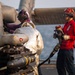 Two military personnel are conducting aircraft servicing operations on what appears to be a naval vessel flight deck over open water. One crew member in a silver/reflective fire-resistant suit and purple helmet is working directly on a military helicopter's weapons/sensor systems, while a second crew member in red shirt and red helmet holds a fueling nozzle/hose. The aircraft visible has mounted ordnance or sensor pods on its stub wings.