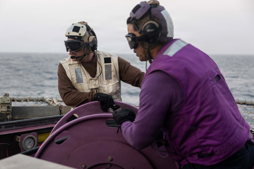 Two military personnel are working together on the deck of a naval vessel at sea. One individual wears a tan/beige flight deck vest with a tactical helmet and communications headset, while the other wears a purple jersey and helmet with goggles, consistent with U.S. Navy flight deck crew roles. They appear to be operating or managing a purple-colored piece of deck equipment, likely a fuel hose reel.