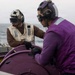 Two military personnel are working together on the deck of a naval vessel at sea. One individual wears a tan/beige flight deck vest with a tactical helmet and communications headset, while the other wears a purple jersey and helmet with goggles, consistent with U.S. Navy flight deck crew roles. They appear to be operating or managing a purple-colored piece of deck equipment, likely a fuel hose reel.