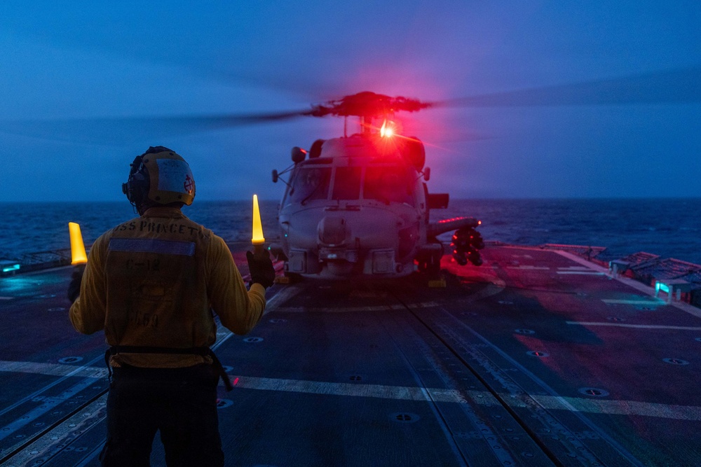 A naval flight deck crew member aboard USS Princeton is guiding a helicopter during twilight operations on a ship's flight deck at sea. The handler is using illuminated yellow wands to direct what appears to be an MH-60 Seahawk helicopter, which has its red navigation/anti-collision lights activated. The scene captures a nighttime or dusk flight deck operation over open ocean.