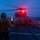 A naval flight deck crew member aboard USS Princeton is guiding a helicopter during twilight operations on a ship's flight deck at sea. The handler is using illuminated yellow wands to direct what appears to be an MH-60 Seahawk helicopter, which has its red navigation/anti-collision lights activated. The scene captures a nighttime or dusk flight deck operation over open ocean.