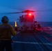 A naval flight deck crew member aboard USS Princeton is guiding a helicopter during twilight operations on a ship's flight deck at sea. The handler is using illuminated yellow wands to direct what appears to be an MH-60 Seahawk helicopter, which has its red navigation/anti-collision lights activated. The scene captures a nighttime or dusk flight deck operation over open ocean.