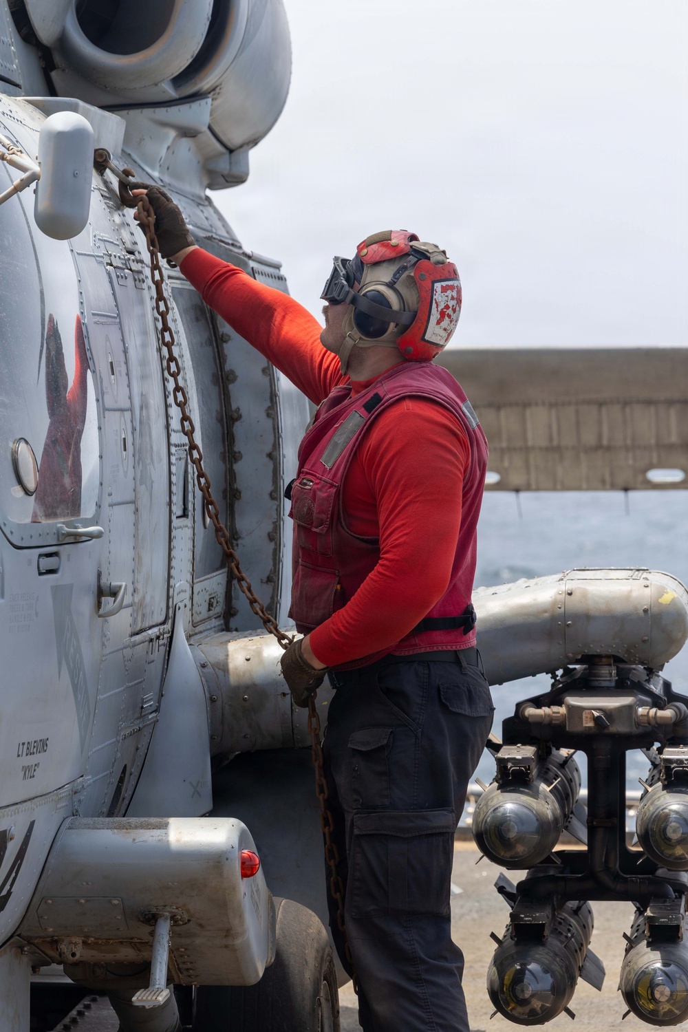 A naval aviation deck crew member wearing red safety gear is securing a military helicopter with a tie-down chain on what appears to be a ship's flight deck. The worker is attaching or checking a chain connected to the aircraft's fuselage, a standard procedure for securing rotary-wing aircraft aboard naval vessels. Text on the aircraft fuselage reads 'LT BLEVINS' and 'KYLE', indicating crew assignment markings.