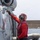 A naval aviation deck crew member wearing red safety gear is securing a military helicopter with a tie-down chain on what appears to be a ship's flight deck. The worker is attaching or checking a chain connected to the aircraft's fuselage, a standard procedure for securing rotary-wing aircraft aboard naval vessels. Text on the aircraft fuselage reads 'LT BLEVINS' and 'KYLE', indicating crew assignment markings.