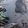 A naval aviation crew member in a green jersey and helmet is servicing a military helicopter numbered 704 on what appears to be a ship's flight deck. The helicopter bears the name 'LCDR SNYDER "MAGIC"' and 'AEAN AGNEW "PUP"' on its fuselage, along with a wolf/fox squadron insignia and a 'RESCUE' triangle marking. A pilot or crew member is visible through the cockpit window.