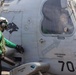 A naval aviation crew member in a green jersey and helmet is servicing a military helicopter numbered 704 on what appears to be a ship's flight deck. The helicopter bears the name 'LCDR SNYDER "MAGIC"' and 'AEAN AGNEW "PUP"' on its fuselage, along with a wolf/fox squadron insignia and a 'RESCUE' triangle marking. A pilot or crew member is visible through the cockpit window.