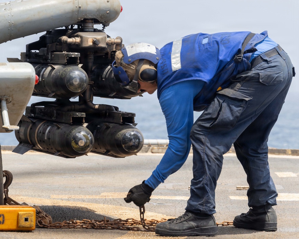 A naval deck crew member in blue coveralls and hearing protection is securing a chain tie-down on a flight deck, likely aboard a naval vessel. The worker is bending over to handle a rusted chain near the landing gear or sensor pod of a military helicopter. The setting appears to be an active ship flight deck with open ocean visible in the background.