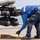 A naval deck crew member in blue coveralls and hearing protection is securing a chain tie-down on a flight deck, likely aboard a naval vessel. The worker is bending over to handle a rusted chain near the landing gear or sensor pod of a military helicopter. The setting appears to be an active ship flight deck with open ocean visible in the background.