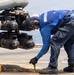 A naval deck crew member in blue coveralls and hearing protection is securing a chain tie-down on a flight deck, likely aboard a naval vessel. The worker is bending over to handle a rusted chain near the landing gear or sensor pod of a military helicopter. The setting appears to be an active ship flight deck with open ocean visible in the background.