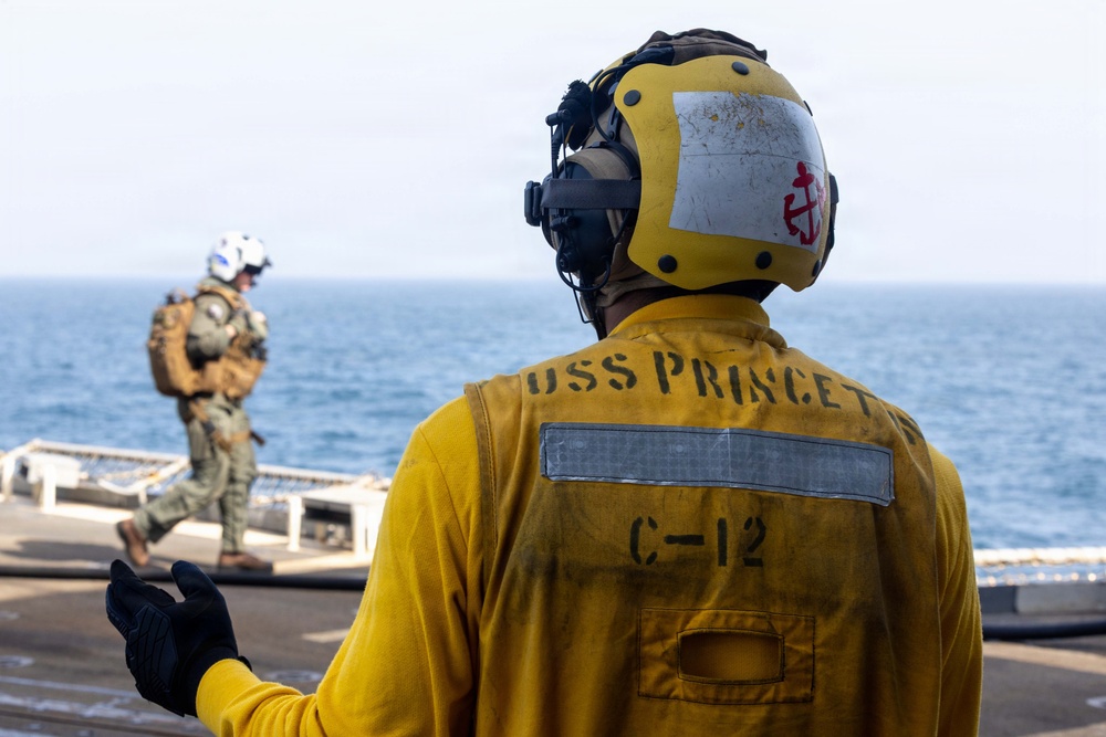 A U.S. Navy sailor in a yellow flight deck jersey marked 'USS PRINCETON' and 'C-12' stands on a ship's flight deck, wearing a yellow helmet with a red anchor insignia and communications headset. In the background, a second personnel member in military tactical gear and a white helmet walks toward the ship's edge with the open ocean visible beyond. The scene depicts active flight deck operations aboard the USS Princeton.