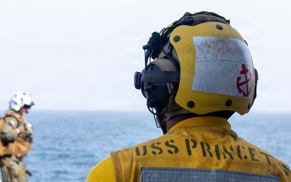 A U.S. Navy sailor in a yellow flight deck jersey marked 'USS PRINCETON' and 'C-12' stands on a ship's flight deck, wearing a yellow helmet with a red anchor insignia and communications headset. In the background, a second personnel member in military tactical gear and a white helmet walks toward the ship's edge with the open ocean visible beyond. The scene depicts active flight deck operations aboard the USS Princeton.