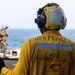 A U.S. Navy sailor in a yellow flight deck jersey marked 'USS PRINCETON' and 'C-12' stands on a ship's flight deck, wearing a yellow helmet with a red anchor insignia and communications headset. In the background, a second personnel member in military tactical gear and a white helmet walks toward the ship's edge with the open ocean visible beyond. The scene depicts active flight deck operations aboard the USS Princeton.