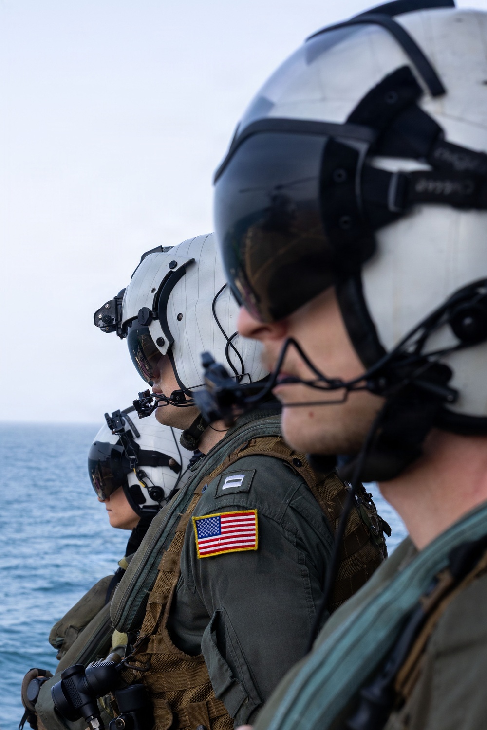 Three military aviators or aircrew members are standing in a line, facing the same direction, with an open ocean visible in the background. They are wearing flight helmets with visors and communication equipment, olive drab flight suits, and tactical gear. The central figure displays a U.S. flag patch on the left shoulder and a rank insignia patch.