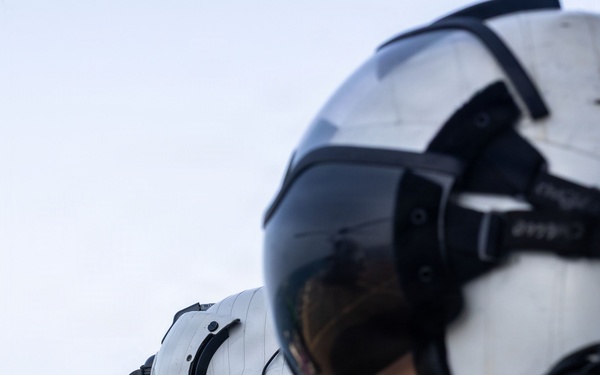 Three military aviators or aircrew members are standing in a line, facing the same direction, with an open ocean visible in the background. They are wearing flight helmets with visors and communication equipment, olive drab flight suits, and tactical gear. The central figure displays a U.S. flag patch on the left shoulder and a rank insignia patch.