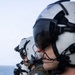 Three military aviators or aircrew members are standing in a line, facing the same direction, with an open ocean visible in the background. They are wearing flight helmets with visors and communication equipment, olive drab flight suits, and tactical gear. The central figure displays a U.S. flag patch on the left shoulder and a rank insignia patch.