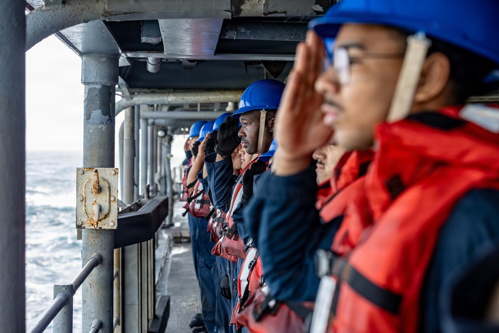 A line of naval personnel standing at attention on the exterior deck of a military vessel at sea, rendering salutes. They are uniformly dressed in navy blue coveralls, blue hard hats, and red inflatable life vests. The image is taken from a low angle looking down the line of sailors, with open ocean visible in the background.