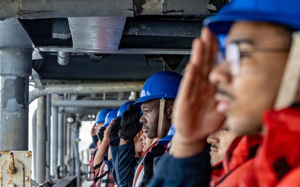 A line of naval personnel standing at attention on the exterior deck of a military vessel at sea, rendering salutes. They are uniformly dressed in navy blue coveralls, blue hard hats, and red inflatable life vests. The image is taken from a low angle looking down the line of sailors, with open ocean visible in the background.