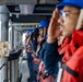 A line of naval personnel standing at attention on the exterior deck of a military vessel at sea, rendering salutes. They are uniformly dressed in navy blue coveralls, blue hard hats, and red inflatable life vests. The image is taken from a low angle looking down the line of sailors, with open ocean visible in the background.