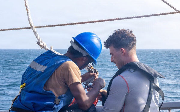 Two individuals are seated on the deck of a naval or maritime vessel at sea. One person wearing a blue hard hat, blue safety vest, and yellow harness appears to be assisting or adjusting equipment on the second person, who is wearing a gray rash guard and an inflatable life vest/harness. The activity appears to be a safety or rescue-related procedure conducted on an open deck with ocean visible in the background.
