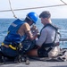 Two individuals are seated on the deck of a naval or maritime vessel at sea. One person wearing a blue hard hat, blue safety vest, and yellow harness appears to be assisting or adjusting equipment on the second person, who is wearing a gray rash guard and an inflatable life vest/harness. The activity appears to be a safety or rescue-related procedure conducted on an open deck with ocean visible in the background.