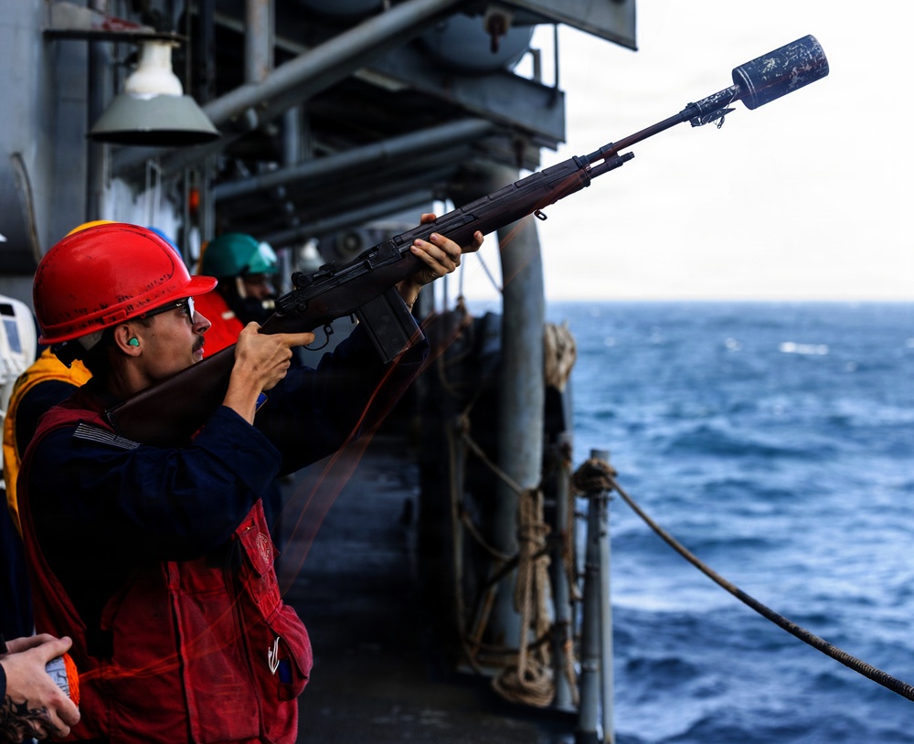 A sailor wearing a red hard hat and hearing protection is firing a line-throwing rifle from the deck of a naval vessel at sea. The rifle has a specialized projectile launcher attached to the muzzle, used for shooting messenger lines between ships during underway replenishment (UNREP) operations. Additional crew members in safety gear are visible in the background.