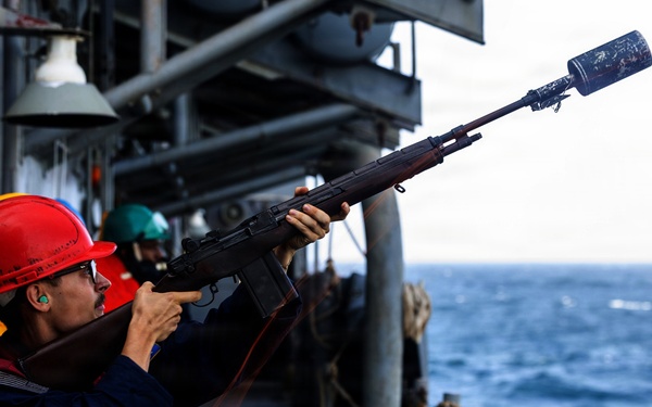 A sailor wearing a red hard hat and hearing protection is firing a line-throwing rifle from the deck of a naval vessel at sea. The rifle has a specialized projectile launcher attached to the muzzle, used for shooting messenger lines between ships during underway replenishment (UNREP) operations. Additional crew members in safety gear are visible in the background.