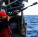 A sailor wearing a red hard hat and hearing protection is firing a line-throwing rifle from the deck of a naval vessel at sea. The rifle has a specialized projectile launcher attached to the muzzle, used for shooting messenger lines between ships during underway replenishment (UNREP) operations. Additional crew members in safety gear are visible in the background.