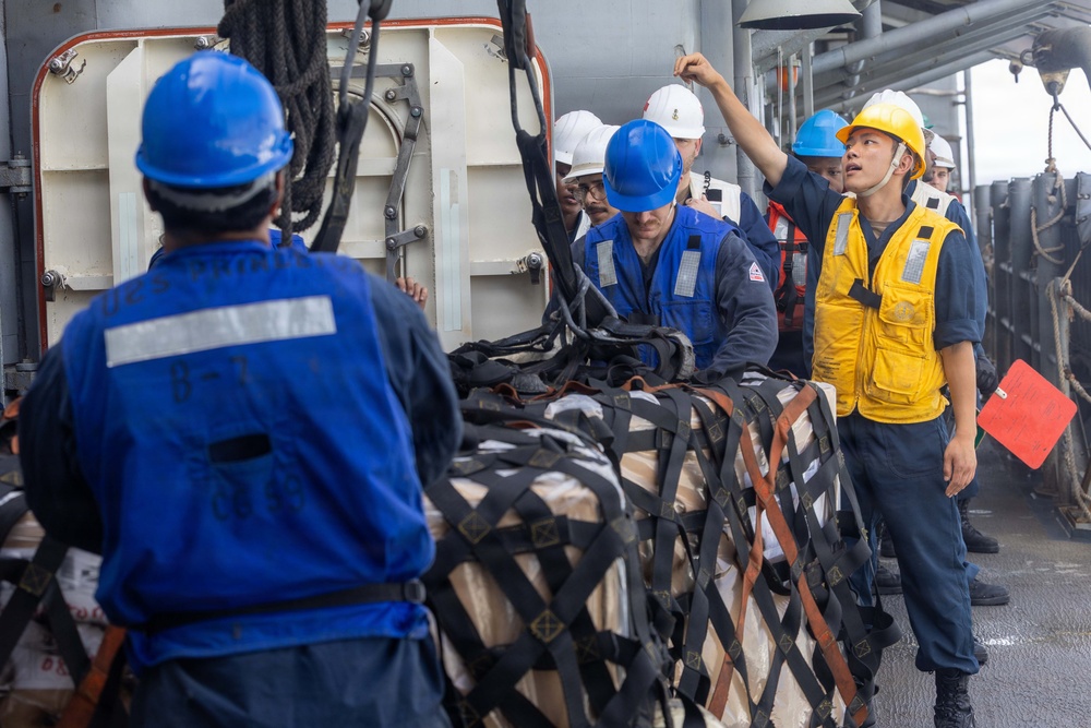 Military or naval personnel are conducting a replenishment-at-sea (RAS) or cargo transfer operation on the deck of a ship. Multiple crew members wearing hard hats and safety vests are managing a netted cargo pallet secured with straps. One crew member in a yellow vest and yellow hard hat appears to be directing or supervising the operation.