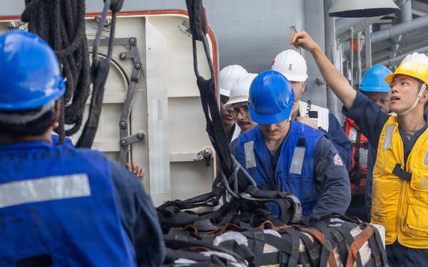 Military or naval personnel are conducting a replenishment-at-sea (RAS) or cargo transfer operation on the deck of a ship. Multiple crew members wearing hard hats and safety vests are managing a netted cargo pallet secured with straps. One crew member in a yellow vest and yellow hard hat appears to be directing or supervising the operation.