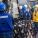 Military or naval personnel are conducting a replenishment-at-sea (RAS) or cargo transfer operation on the deck of a ship. Multiple crew members wearing hard hats and safety vests are managing a netted cargo pallet secured with straps. One crew member in a yellow vest and yellow hard hat appears to be directing or supervising the operation.