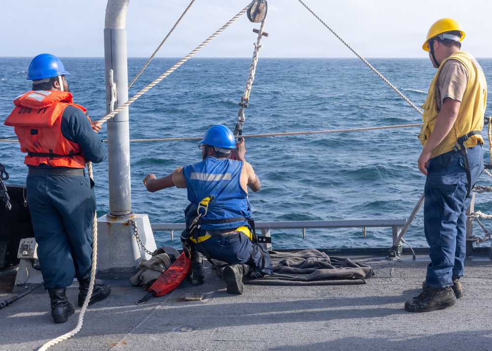 Three personnel are working on the deck of a naval vessel at sea. Two workers wearing blue hard hats and safety vests are actively handling rigging equipment near a pulley system mounted on a vertical pole, while a third person in a yellow hard hat and yellow shirt observes. The operation appears to involve rope and block-and-tackle rigging work over the ship's side.
