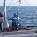 Three personnel are working on the deck of a naval vessel at sea. Two workers wearing blue hard hats and safety vests are actively handling rigging equipment near a pulley system mounted on a vertical pole, while a third person in a yellow hard hat and yellow shirt observes. The operation appears to involve rope and block-and-tackle rigging work over the ship's side.