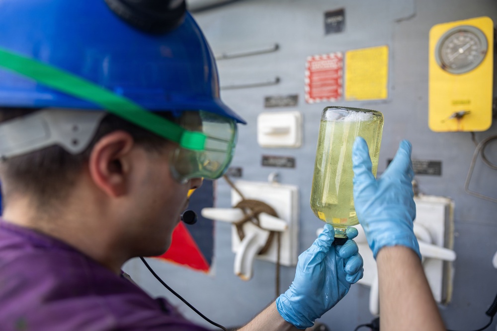 A worker wearing a blue hard hat, green safety goggles, and blue nitrile gloves is examining a glass container filled with a yellow-green liquid in an industrial or marine environment. The person appears to be conducting a visual inspection or quality check of the fluid sample. The background shows a gray wall with mounted gauges, warning labels, and equipment panels.