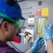 A worker wearing a blue hard hat, green safety goggles, and blue nitrile gloves is examining a glass container filled with a yellow-green liquid in an industrial or marine environment. The person appears to be conducting a visual inspection or quality check of the fluid sample. The background shows a gray wall with mounted gauges, warning labels, and equipment panels.