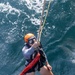 A rescue swimmer or combat swimmer is being hoisted out of open ocean water via a rope/hoist line. The individual is wearing full water rescue gear including an orange helmet, dive goggles, a harness, inflated life vest, and swim fins. They are giving a thumbs-up signal, indicating they are ready or okay during what appears to be a military or coast guard rescue training exercise.