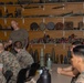 A military instructor in a olive drab t-shirt is conducting a briefing or training session for a group of uniformed Marines in what appears to be an ordnance or explosive ordnance disposal (EOD) training room. The walls are lined with shelves displaying a wide variety of inert munitions, projectiles, mines, rockets, and ordnance items used as training aids. The students are seated at tables facing the instructor in a classroom-style setting.