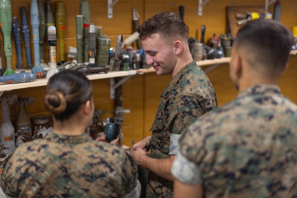 Three military personnel in MARPAT camouflage uniforms are gathered in what appears to be an ordnance or EOD (Explosive Ordnance Disposal) training room or armory. One female service member is holding a dark-colored hand grenade while a male service member facing the camera appears to be examining or discussing an item. The shelves behind them display a large collection of inert training munitions, rockets, and ordnance items.